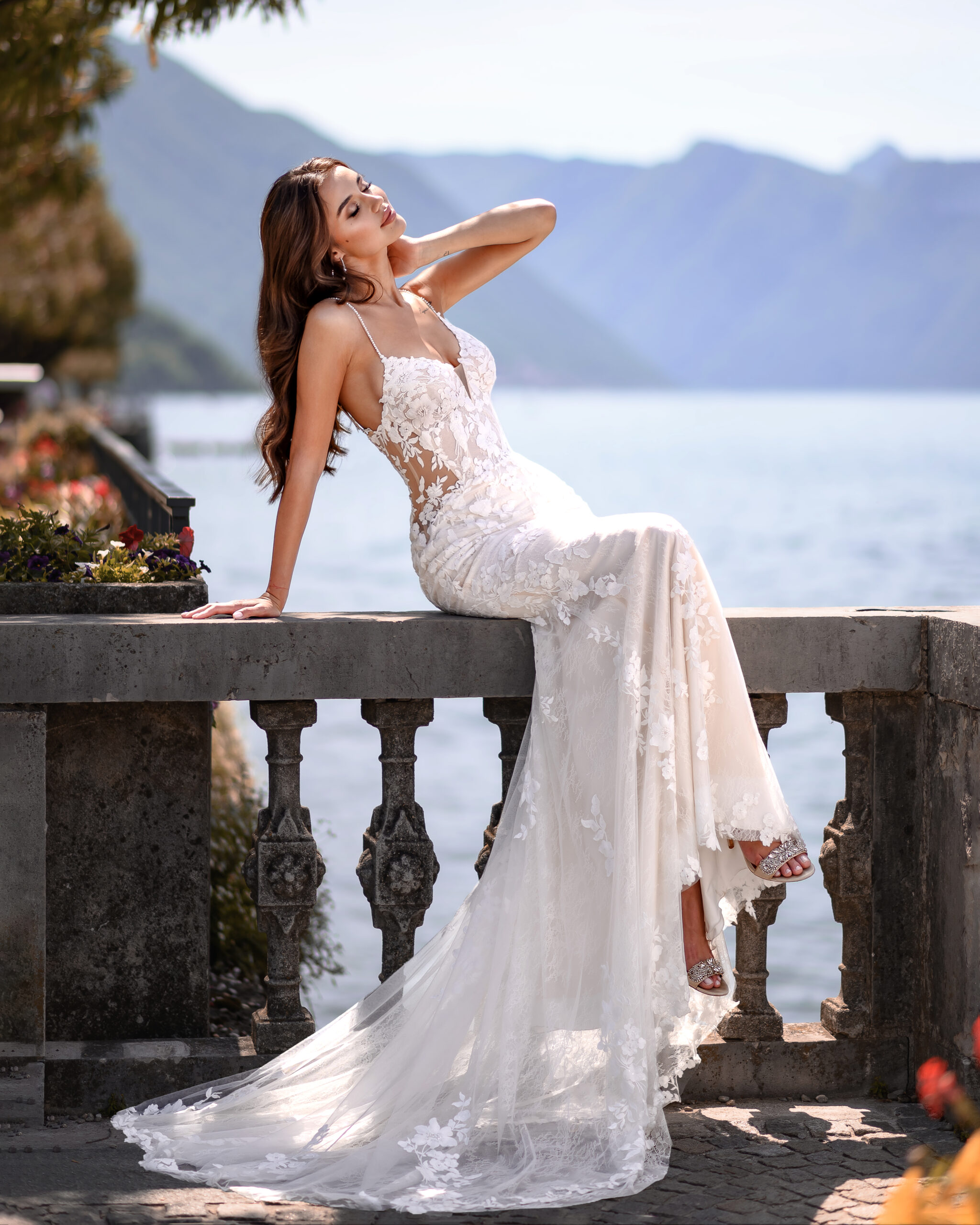 Eine Frau in einem weißen Spitzen-Brautkleid sitzt auf einer Steinbrüstung am Ufer eines Sees.